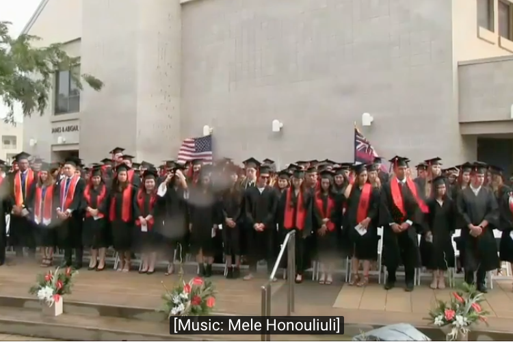 Graduates on stage at commencement with closed captioning stating music is playing.