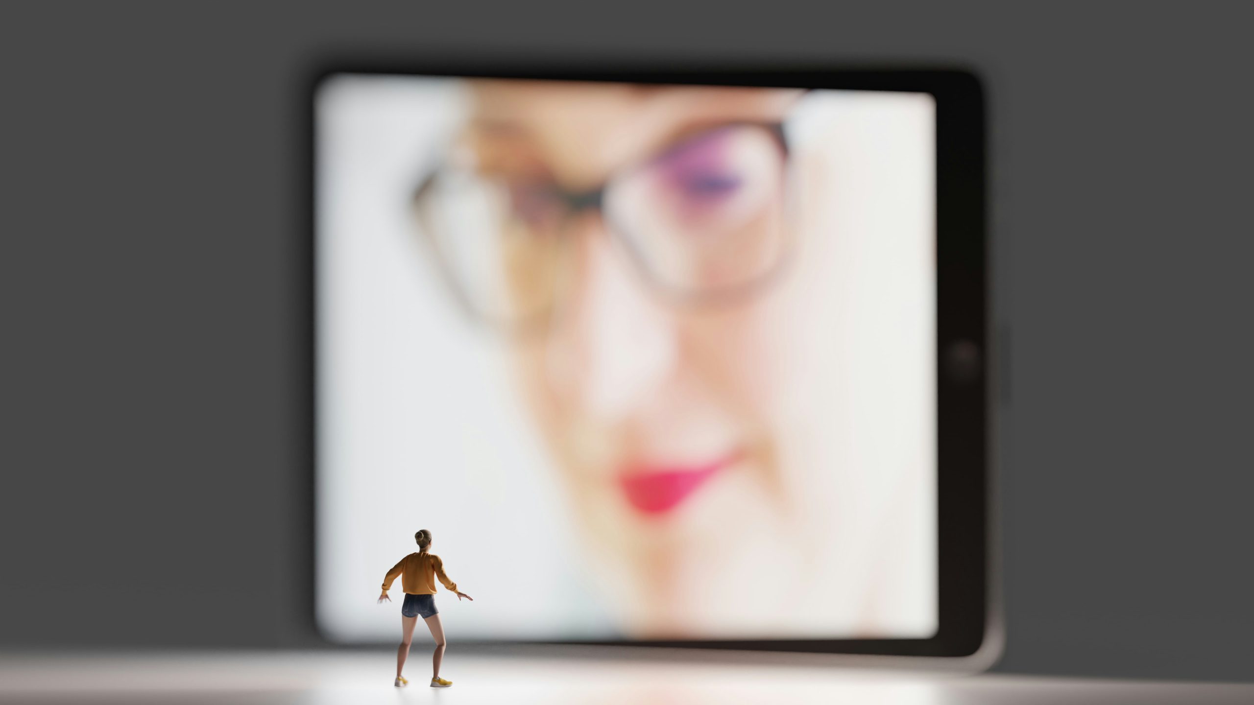 A tiny action figure looks up at a blurred female face that fills a computer monitor.