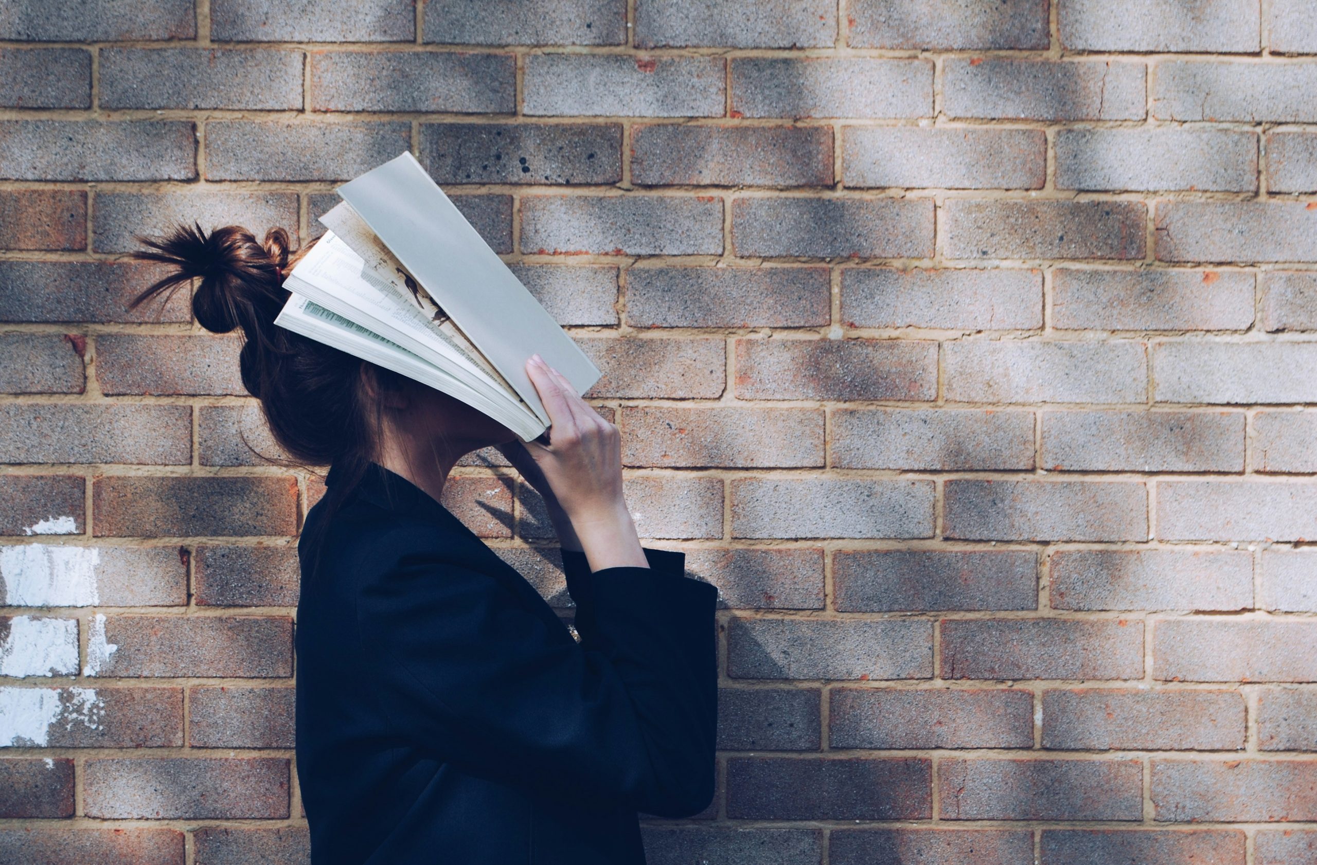 Woman covers her face with a textbook with brick wall as backdrop.
