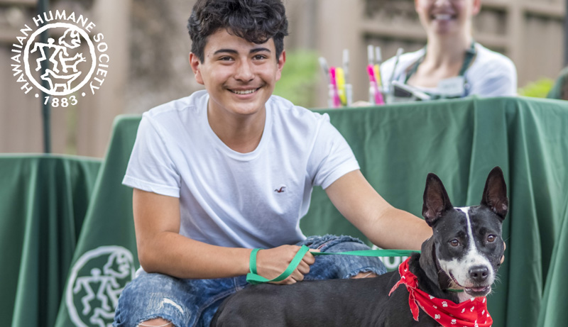 A boy and his dog squatting in front of a Hawaiian Humane Society outreach table.