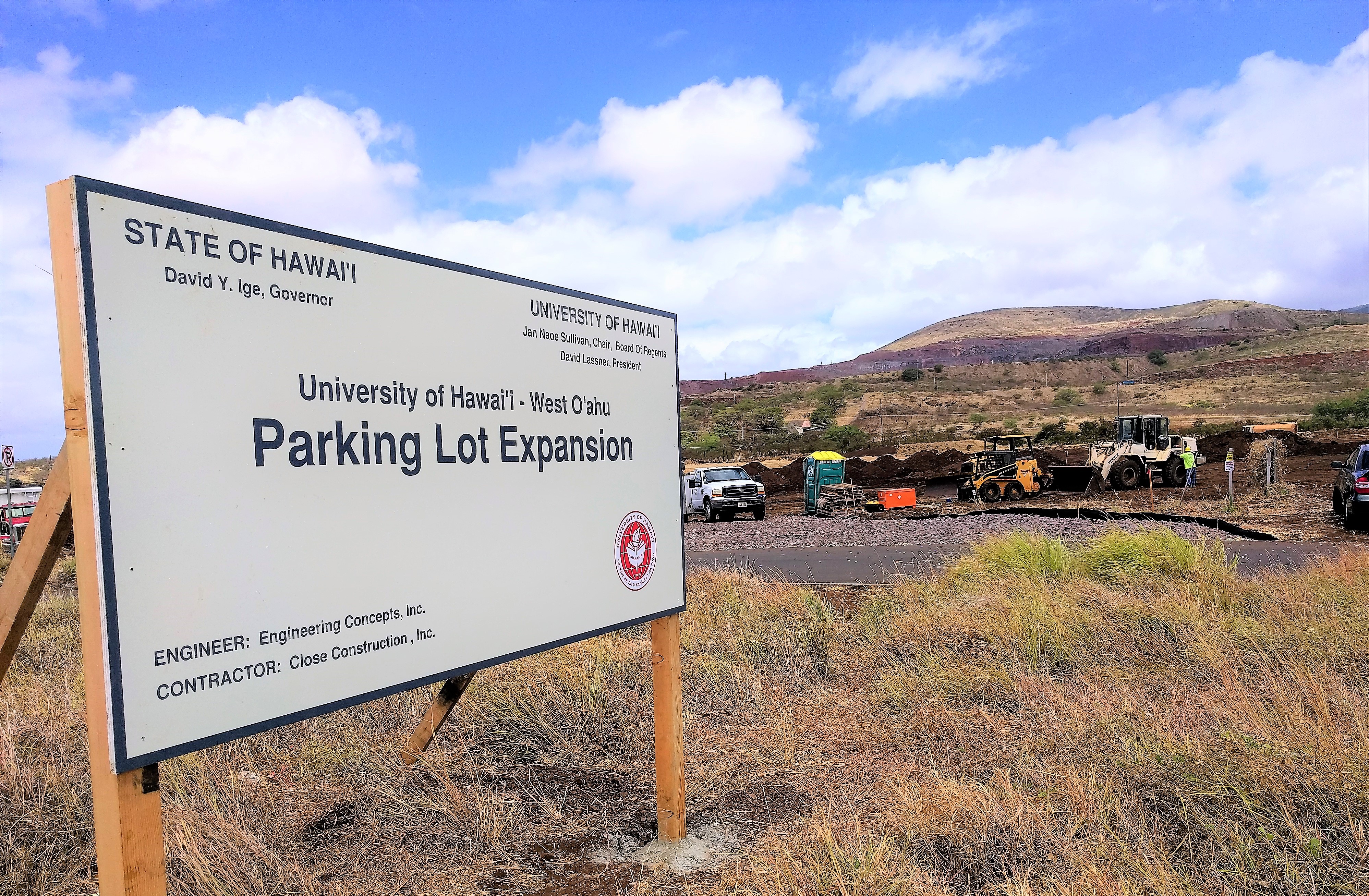 Photo of sign announcing parking lot expansion in foreground, vehicles and worksite in background