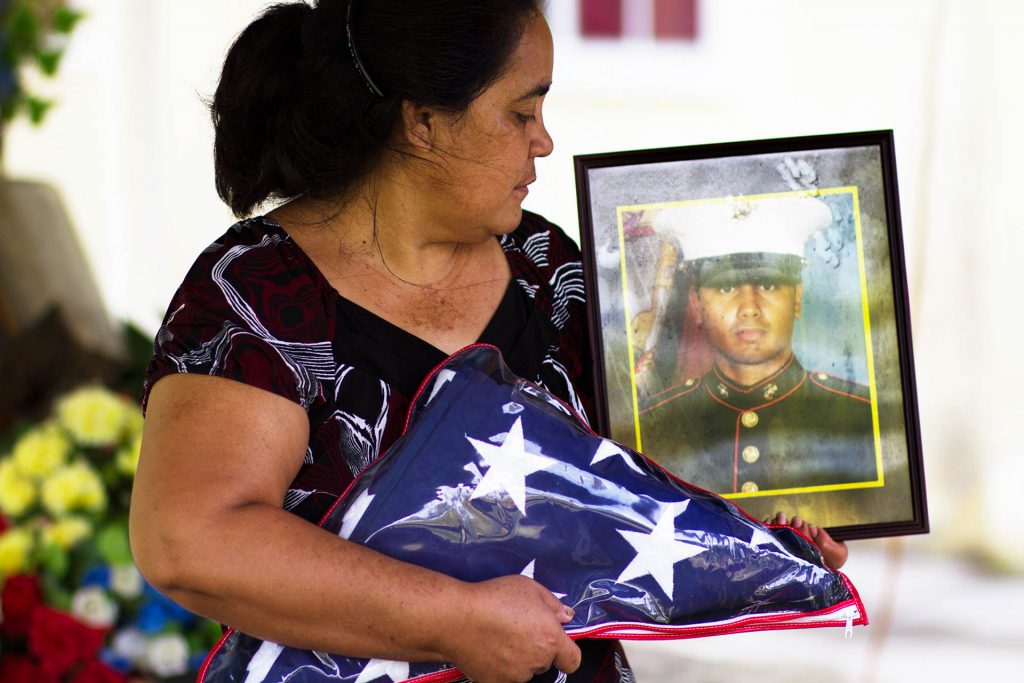 Photo of woman holding a folded flag with photo of deceased soldier in the background