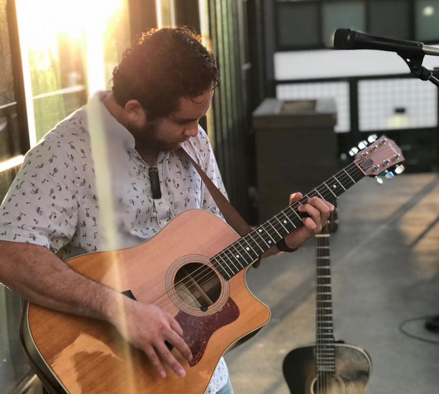 Shot of Danny Carvalho standing against some windows at sunset while playing a guitar. The sun is reflected off of the windows