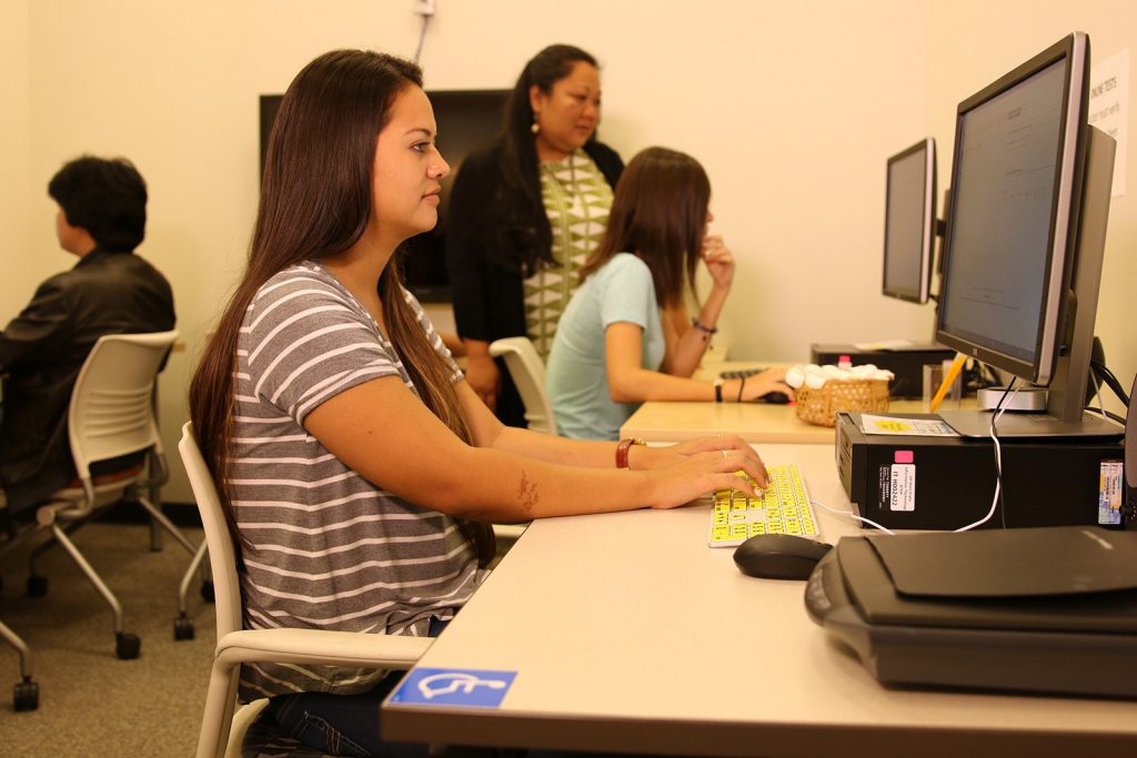 Photo of two people taking tests at desktop computers with a test proctor in the background
