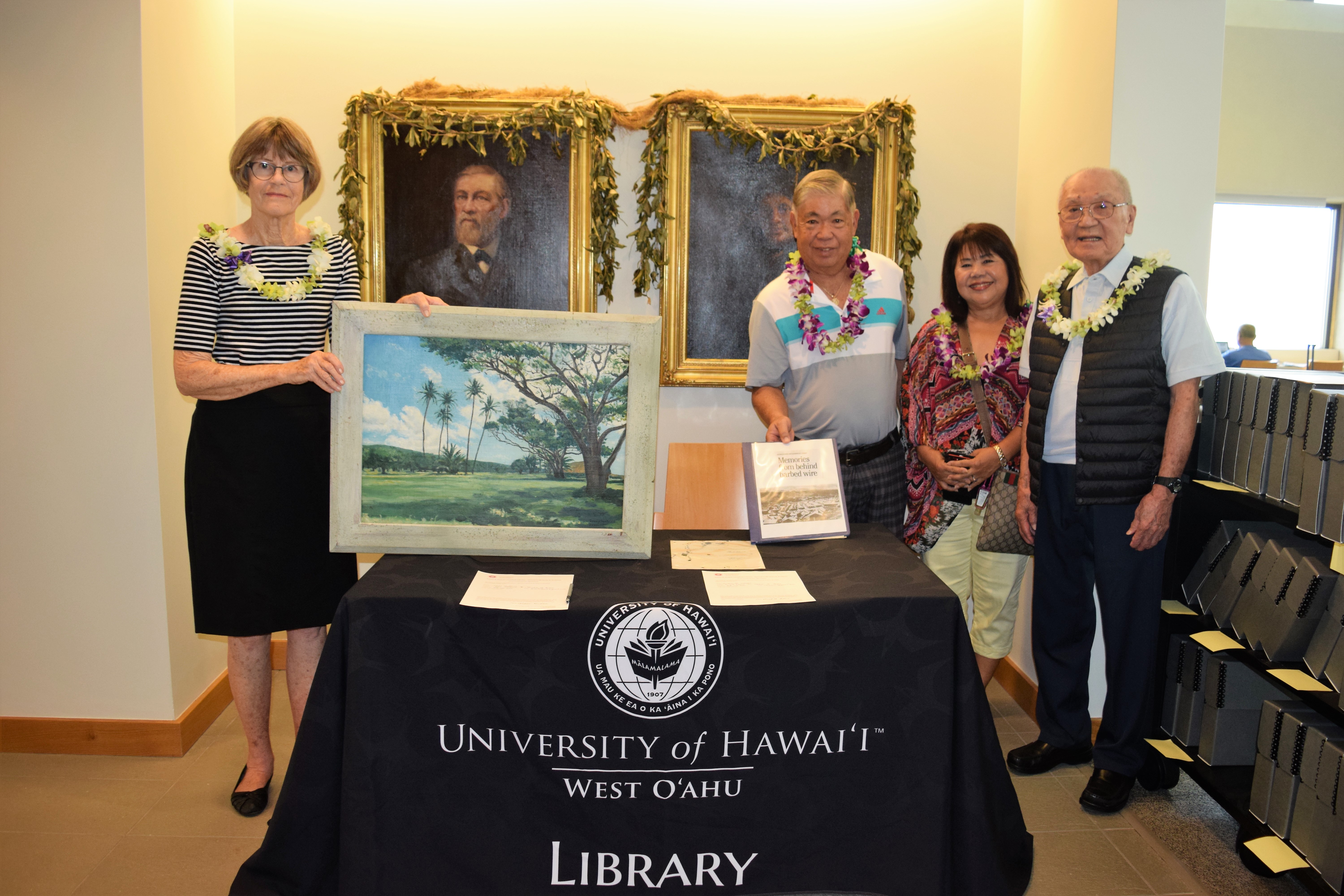 photo of four people standing behind a table. Woman on the left, Letitia Hickson is holding up a painting she donated, while to the right of her Ronald Ikehara holds up a notebook containing letters from POWs
