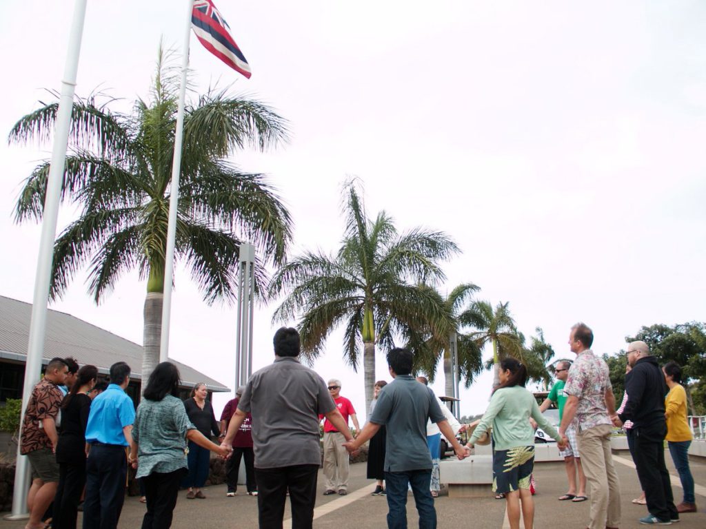 photo of people standing in a circle, holding hands, with Hawaiian flag flying above them