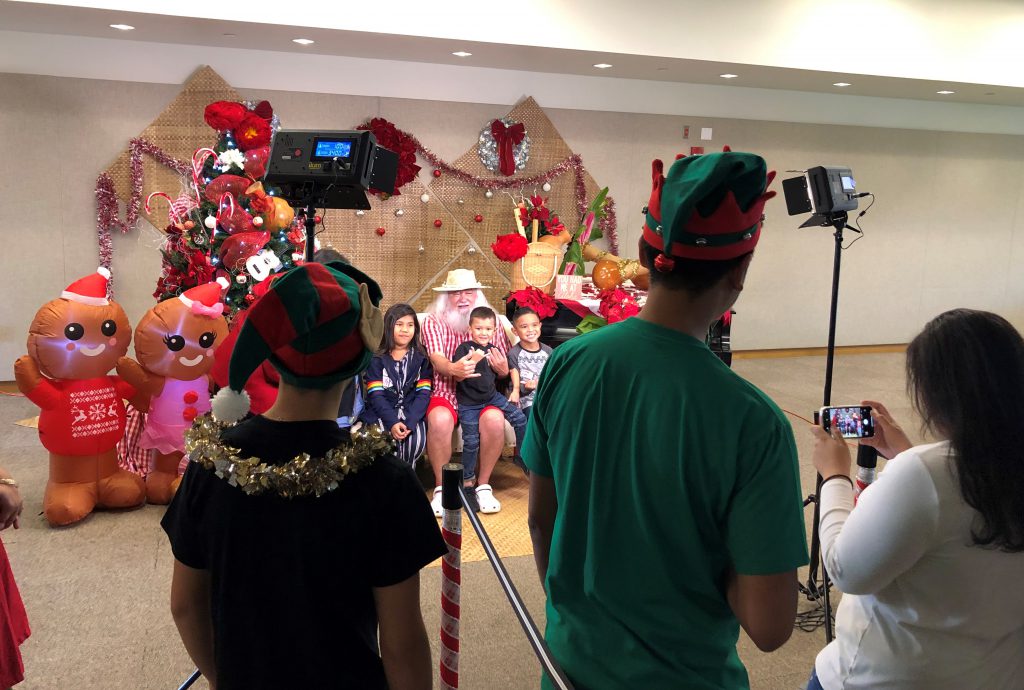 Photo of children sitting with Santa in shorts being photographed and watched by other people