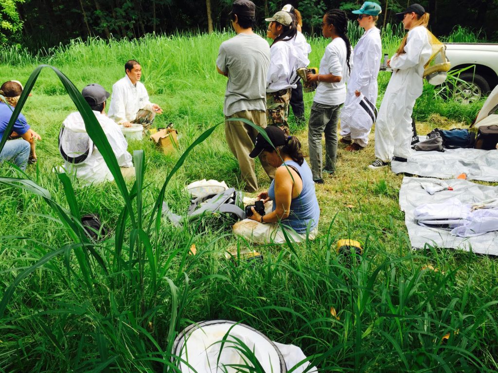 photo of an instructor and students outdoors. Students are mainly standing, dressed in Bee Keeper suits and an instructor is kneeling, talking to them.