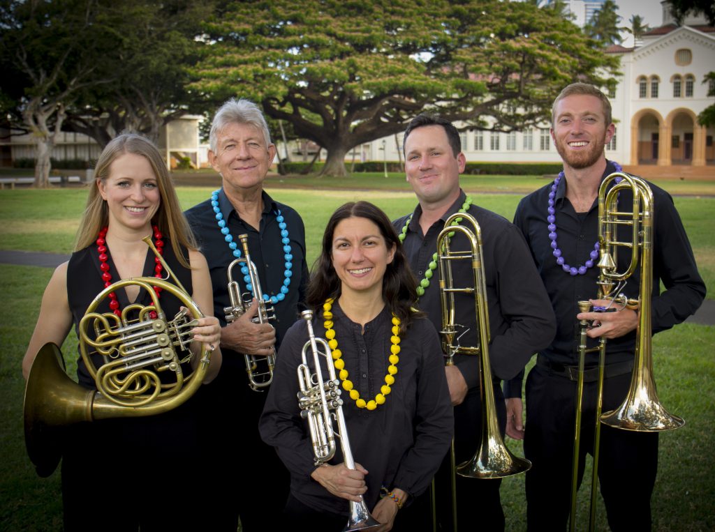 Photo of two women and three men standing on a lawn in front of trees and a building. Each are holding a brass musical instrument