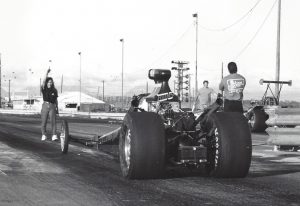 Black and white photo of a dragster at Hawaii Raceway Park. Three people are in the photo, including a woman gesturing with her arm as she walks toward the dragster