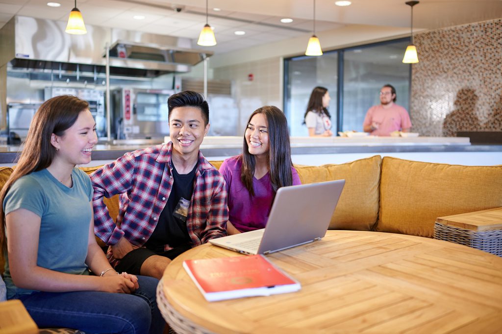 Photo of three students sitting at a table, apparently talking about an assignment or whatever is on the screen of the laptop in front of them. In the background are two students standing and talking.