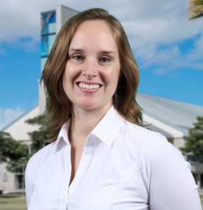 Portrait photo of Dr. Jennifer Byrnes. She is wearing a white blouse and smiling. The backdrop is the UH West Oahu library building.
