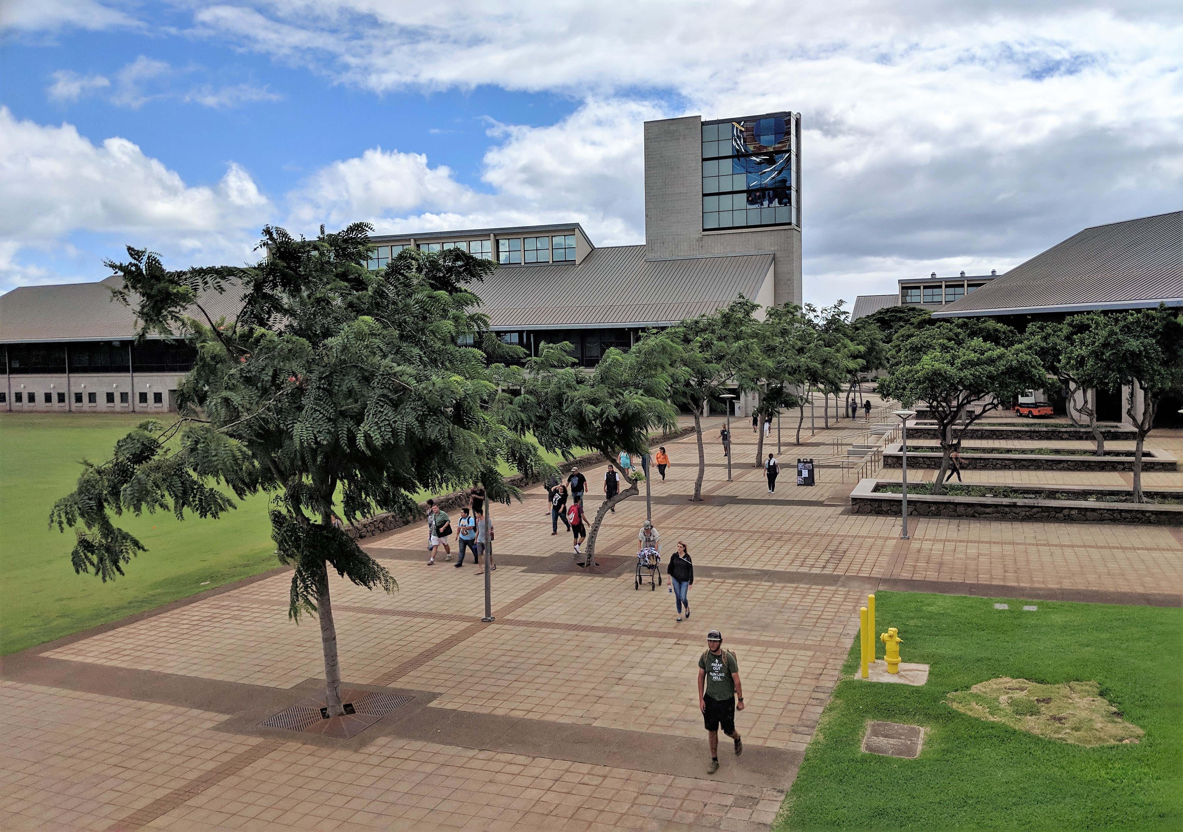 photo of the UH West Oahu campus looking from the Campus Center down at the broad walkway fronting the Campus Center Plaza. Students are walking and trees line the concrete walkway. In the background is the library building