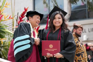 Photo of graduation with Chancellor Benham on left smiling and shaking the hand of graduate Chael Kekona, who is holding a diploma holder