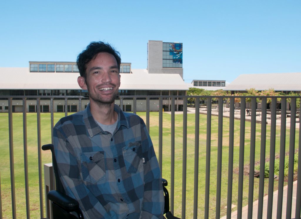 Photo of a man sitting in a wheelchair. Behind him is a metal railing and beyond that is the UH West Oahu campus - You can see portions of the Great Lawn, the Library, Campus Center and the Laboratory building