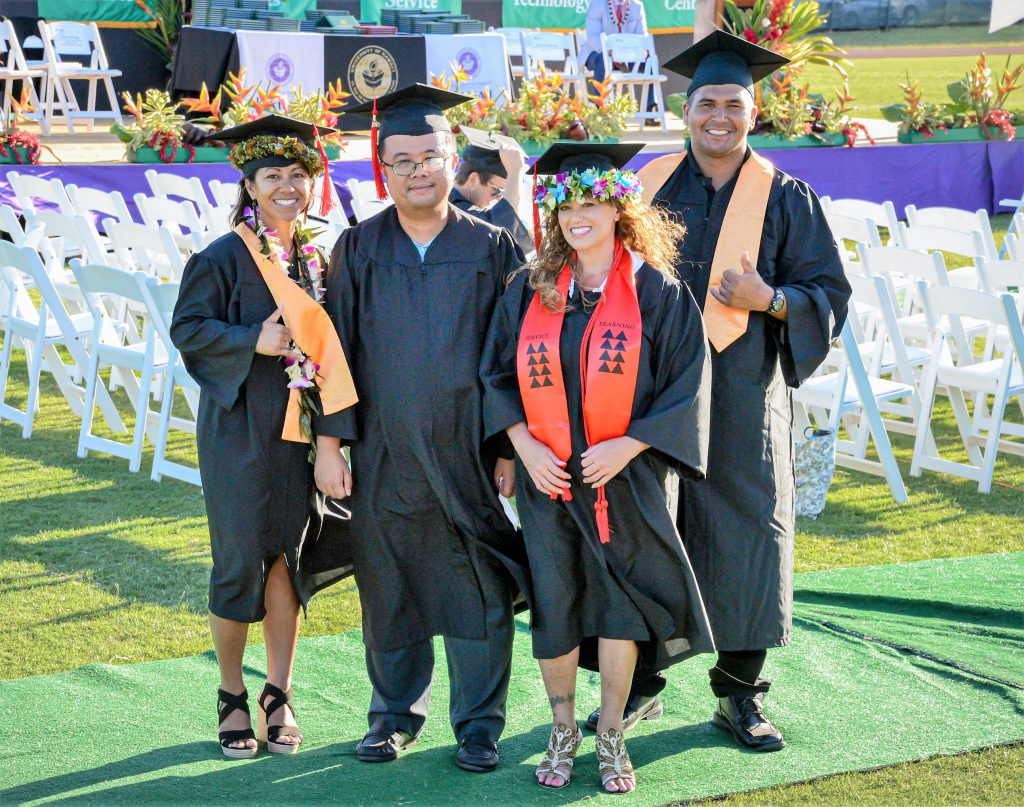 photo of four people in graduation caps and gowns standing on a grassy lawn with white folding chairs and a stage in the background. There are two men and two women. One of the men is flashing a shaka sign