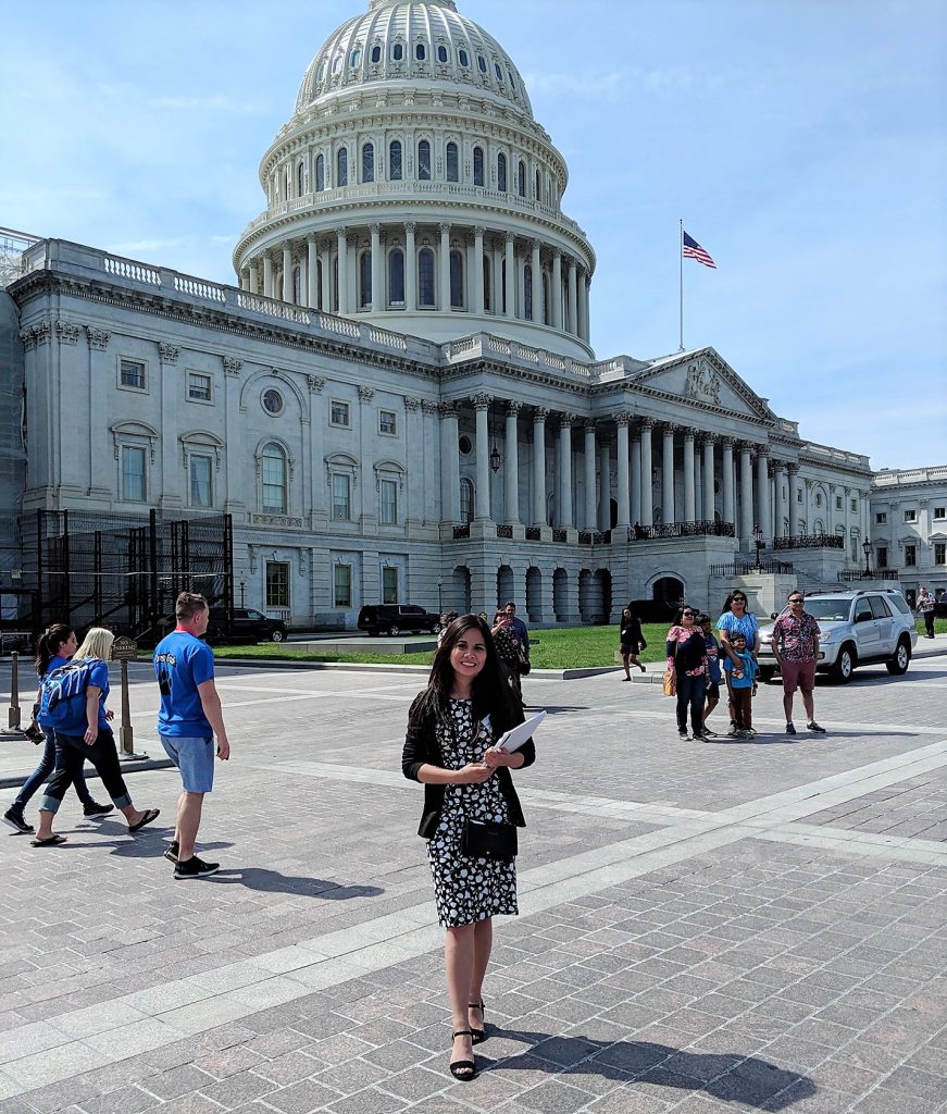 Photo of a woman holding some papers posed in front of the U.S. Capitol. It is a bright, sunny day and in the background tourists are walking around