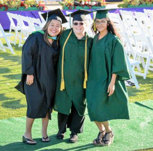 Photo of three female graduating students. One is wearing a black cap and gown and the other two are wearing green cap and gowns. There are white folding chairs in the background. They are standing on green astroturf
