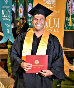Photo of a man walking down a wood ramp holding a diploma while dressed in a cap and gown. He is smiling