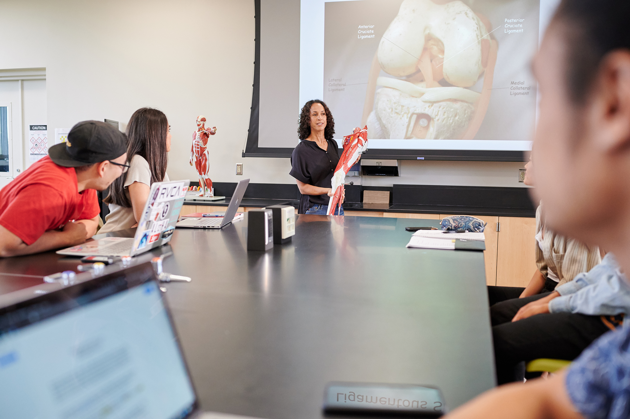 Photo of a professor in a classroom and students listening to her. The professor is holding what appears to be a model of the human leg and on the screen behind her is a diagram of a human leg