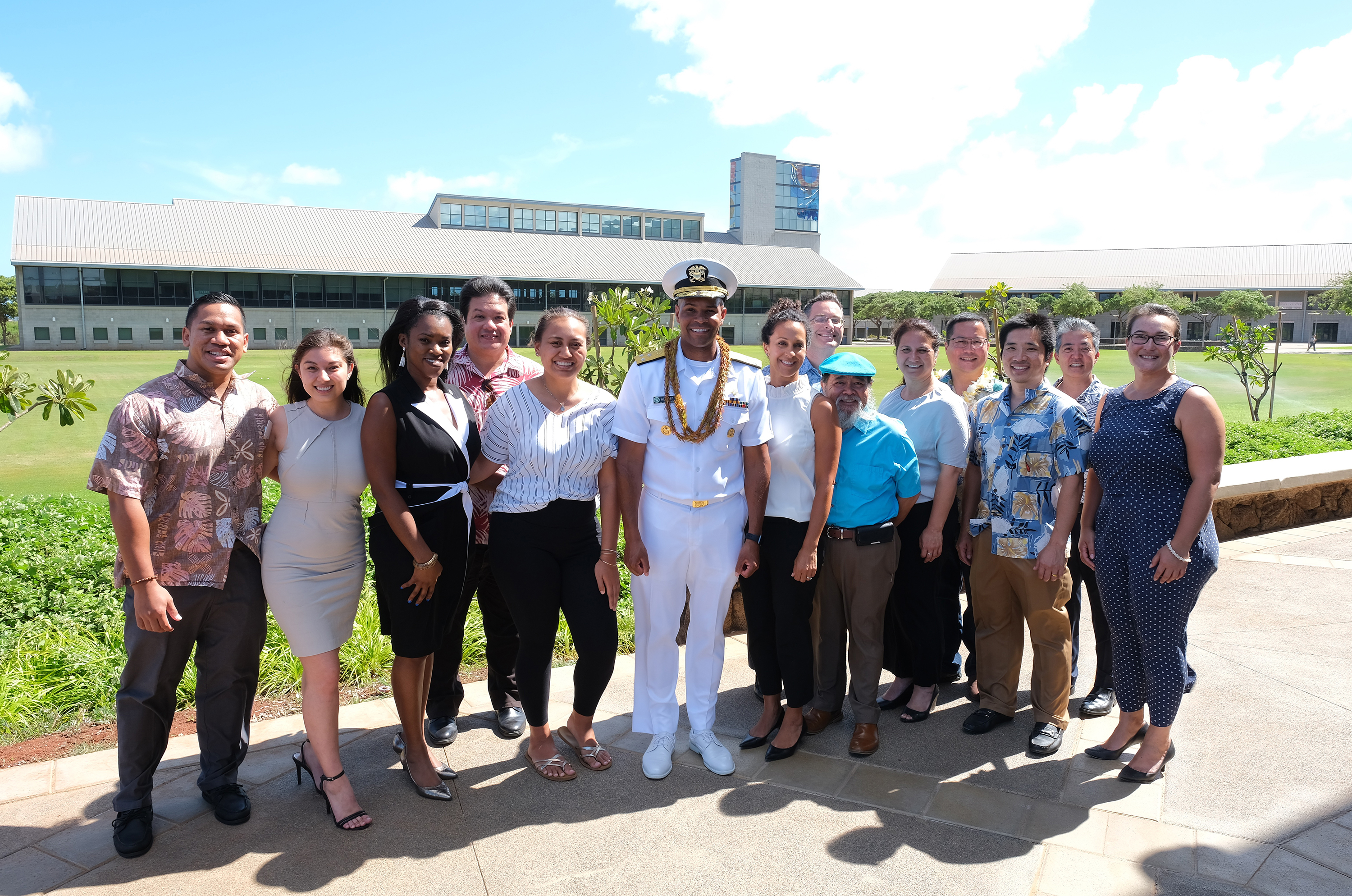 Photo of a group of people with a man in a white uniform standing at center. In the background is the UH West Oahu Great Lawn and the James & Abigail Campbell Library