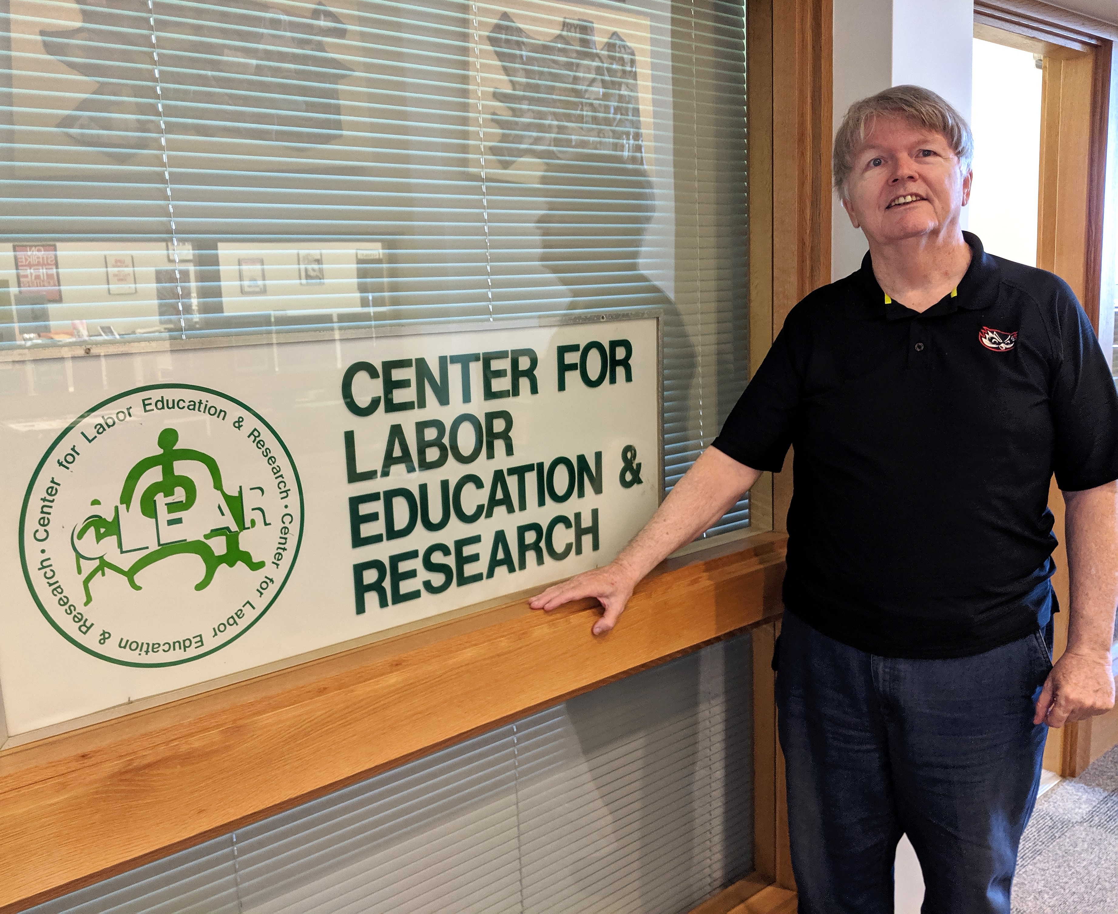 Photo of a man in a black polo shirt standing next to a window to an office with the shades drawn closed. In the window is a sign that says Center for Labor Education and Research