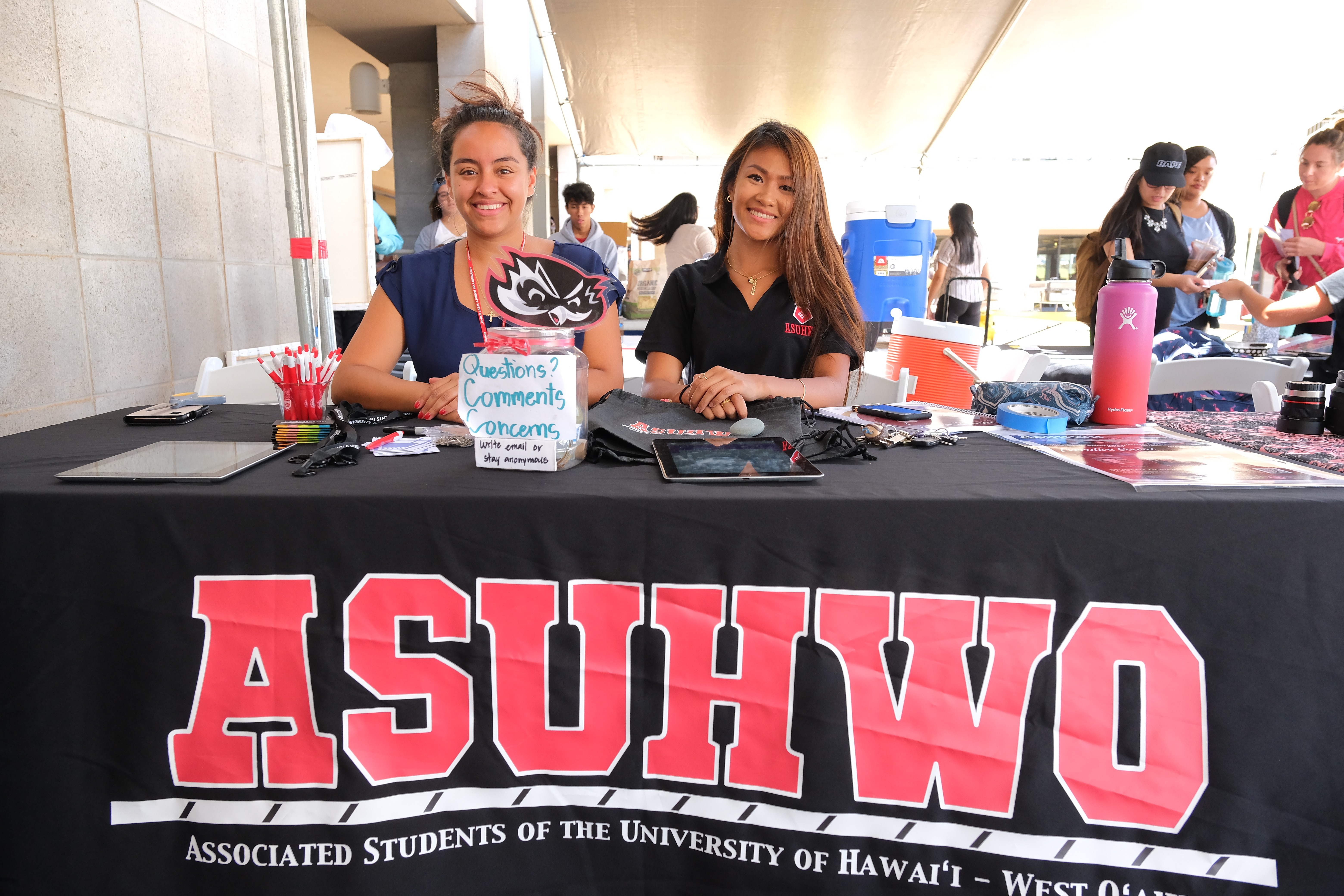 Two female students smiling, sitting at a table to promote the Associated Students of the University of Hawaii-West Oahu.
