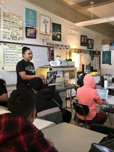 Naturalee ʻIlima Puou standing at the front of a classroom and speaking to students, who are sitting at their desks.