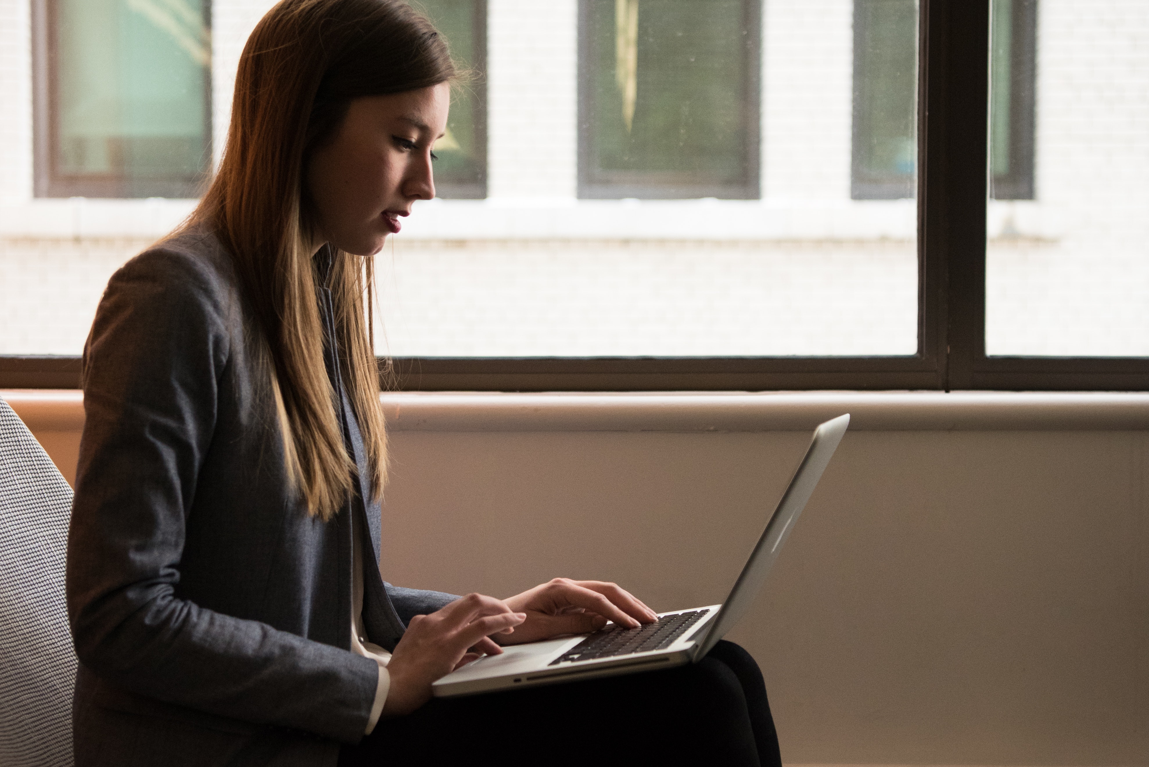 Woman sitting by a window and typing on a laptop.