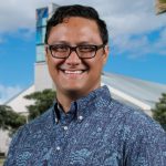 Portrait photo of Dr. Kamuela Yong. He is wearing a blue on blue aloha shirt. The background is the library building