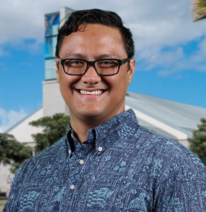 Portrait photo of Dr. Kamuela Yong. He is wearing a blue on blue aloha shirt. The background is the library building