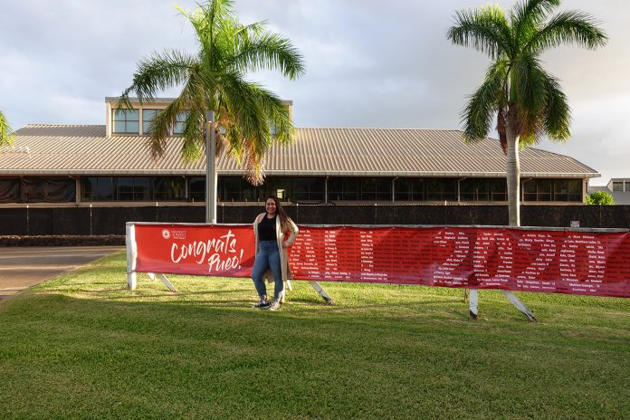 Woman smiling and standing in front of a commencement banner on UH West Oʻahu campus.