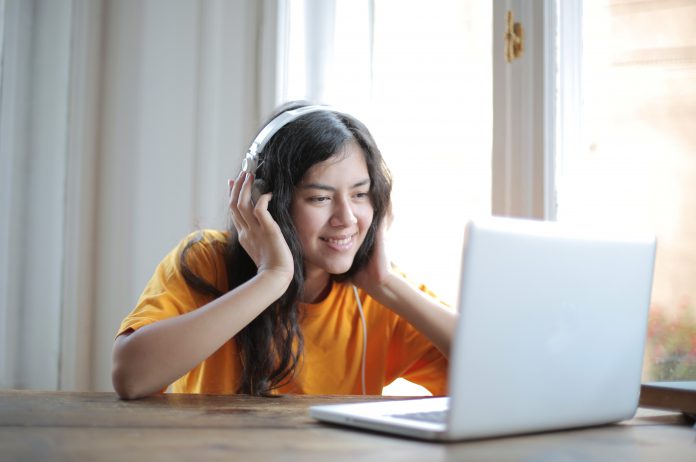 Woman in orange shirt and headphones, sitting at a table and looking at her laptop.