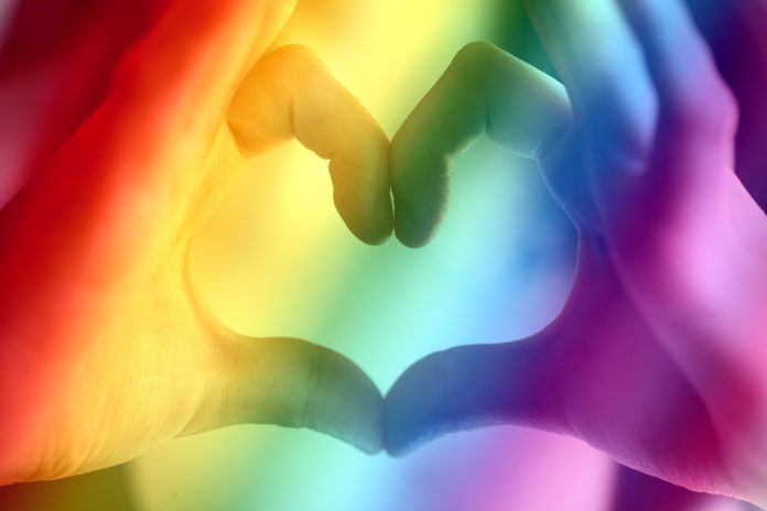 Fingers making the shape of a heart with rainbow colors in the foreground.