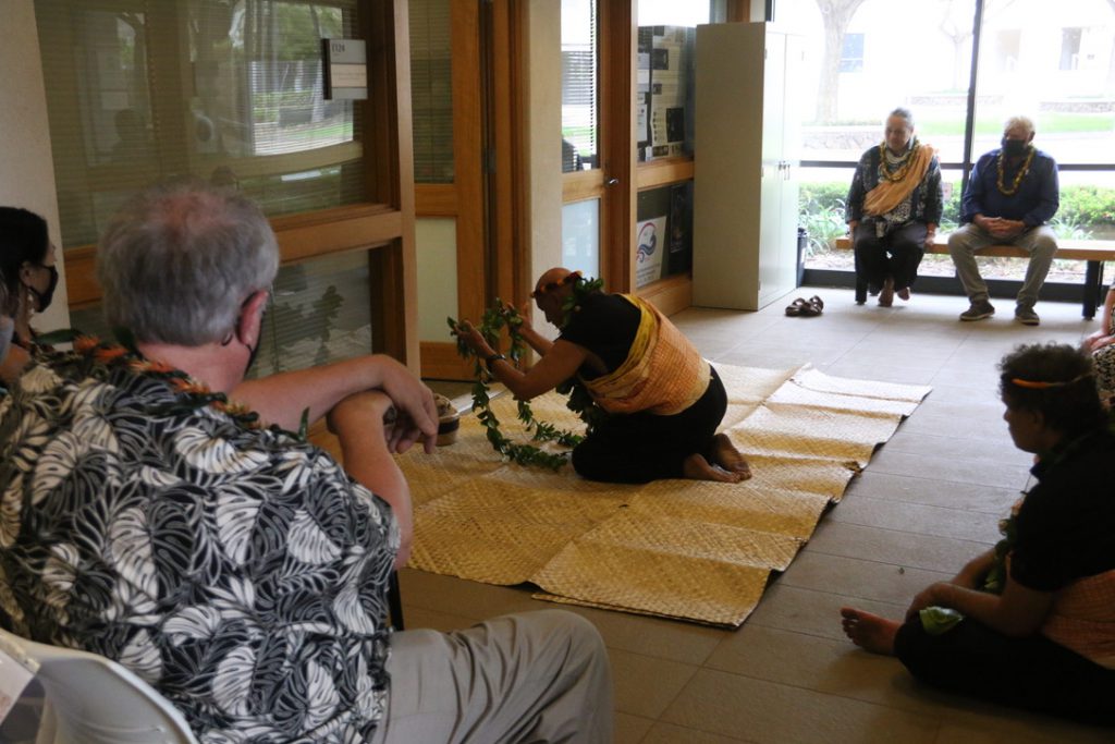 A man kneeling and holding a maile lei with a surrounding audience observing.