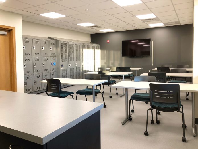 A room with tables, chairs, a wall-mounted screen, and lockers.