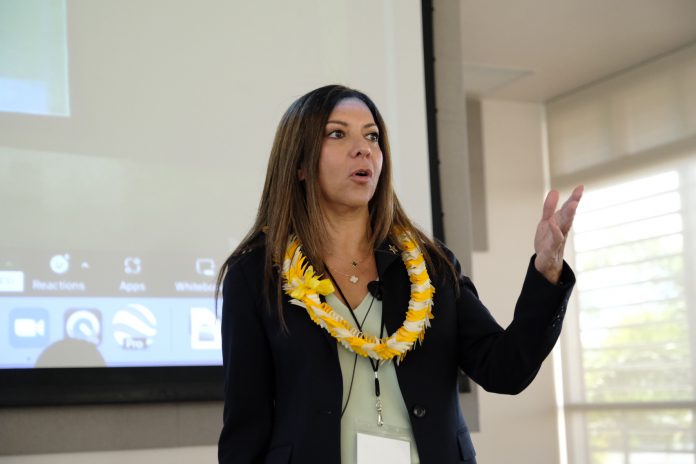 A woman wearing a lei and speaking in front of a large screen.