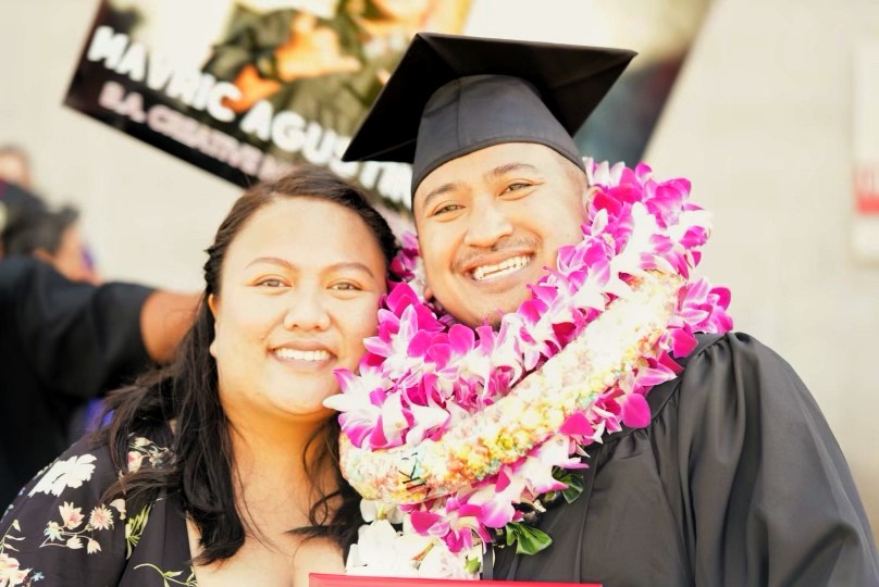 A married couple at the husbandʻs graduation ceremony. The husband is wearing a cap, gown, and many lei.