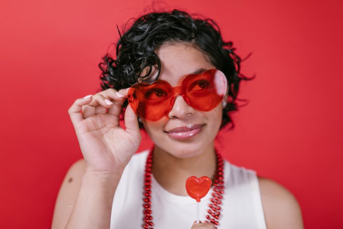 A woman wearing red, heart-shaped glasses and holding a red heart lollipop.