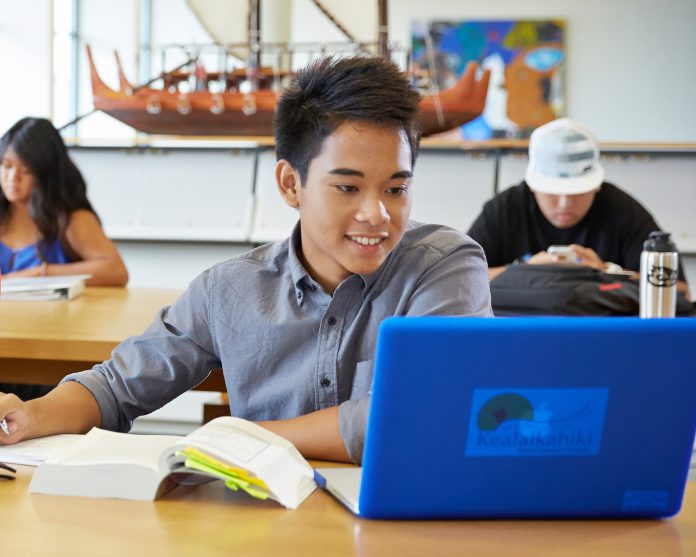 A student looking at his blue laptop with two other students studying in the background in the room.