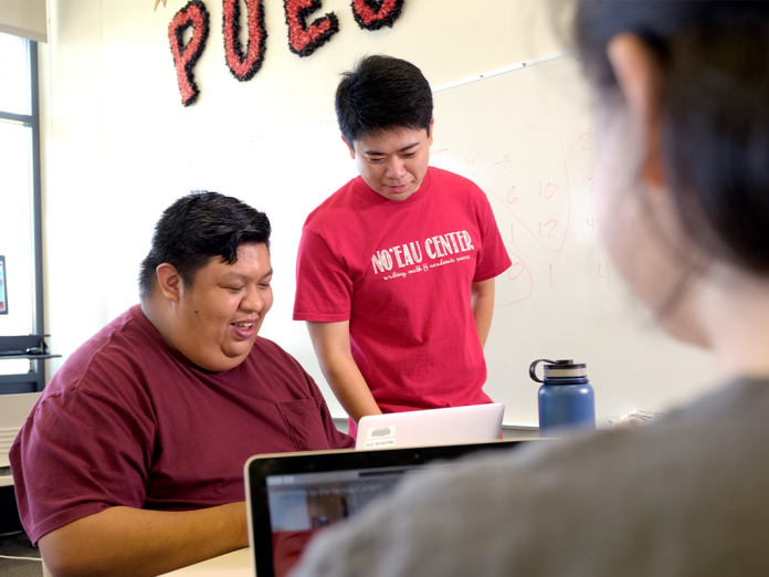 A student sitting at a table with a laptop, with a tutor standing next to him. Both are smiling and looking at the studentʻs laptop.