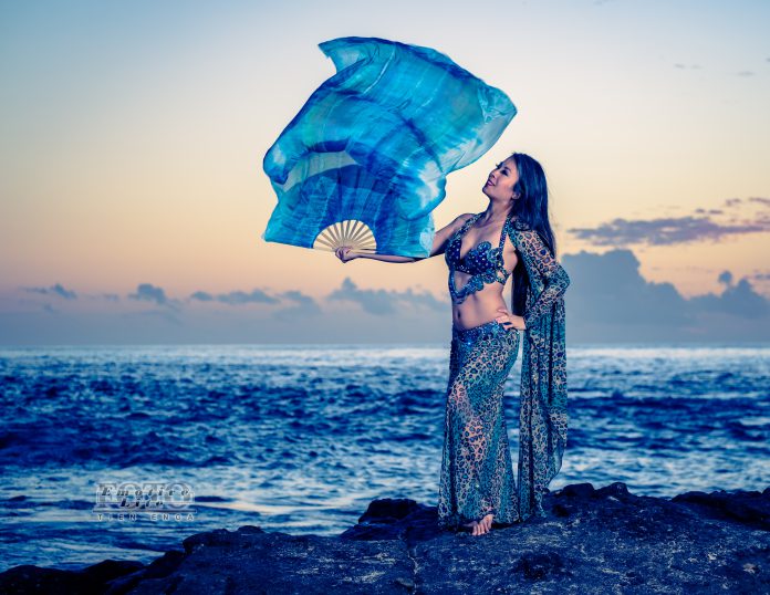 A woman wearing belly dancing attire and standing on rocks with the ocean and a sunset in the background.