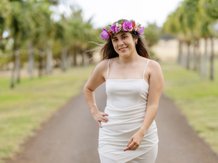 Woman wearing a head lei and white dress, and walking down a path with trees behind her.