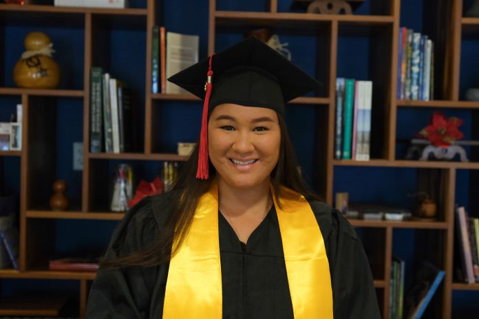 A woman wearing a graduation cap and gown and standing in front of a bookshelf.