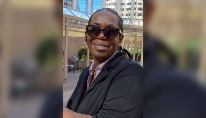Justice Shorter, a Black woman, wears a lavender shirt, a navy blue blazer, and sunglasses. She is standing in front of a modern building and her hair is styled in straight back cornrows.