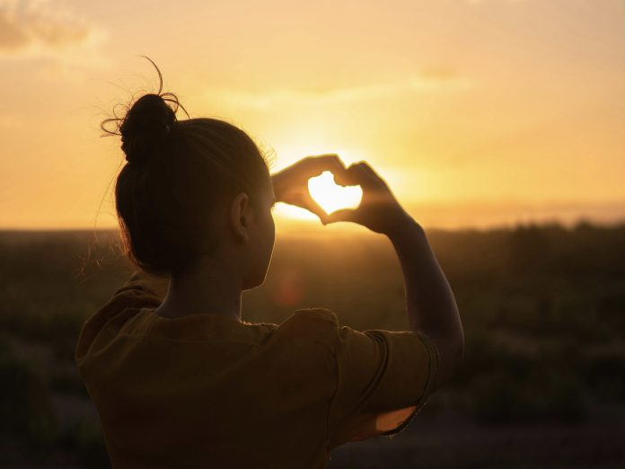 Silhouette of a woman making the shape of a heart with her hands with a sunset in the background.