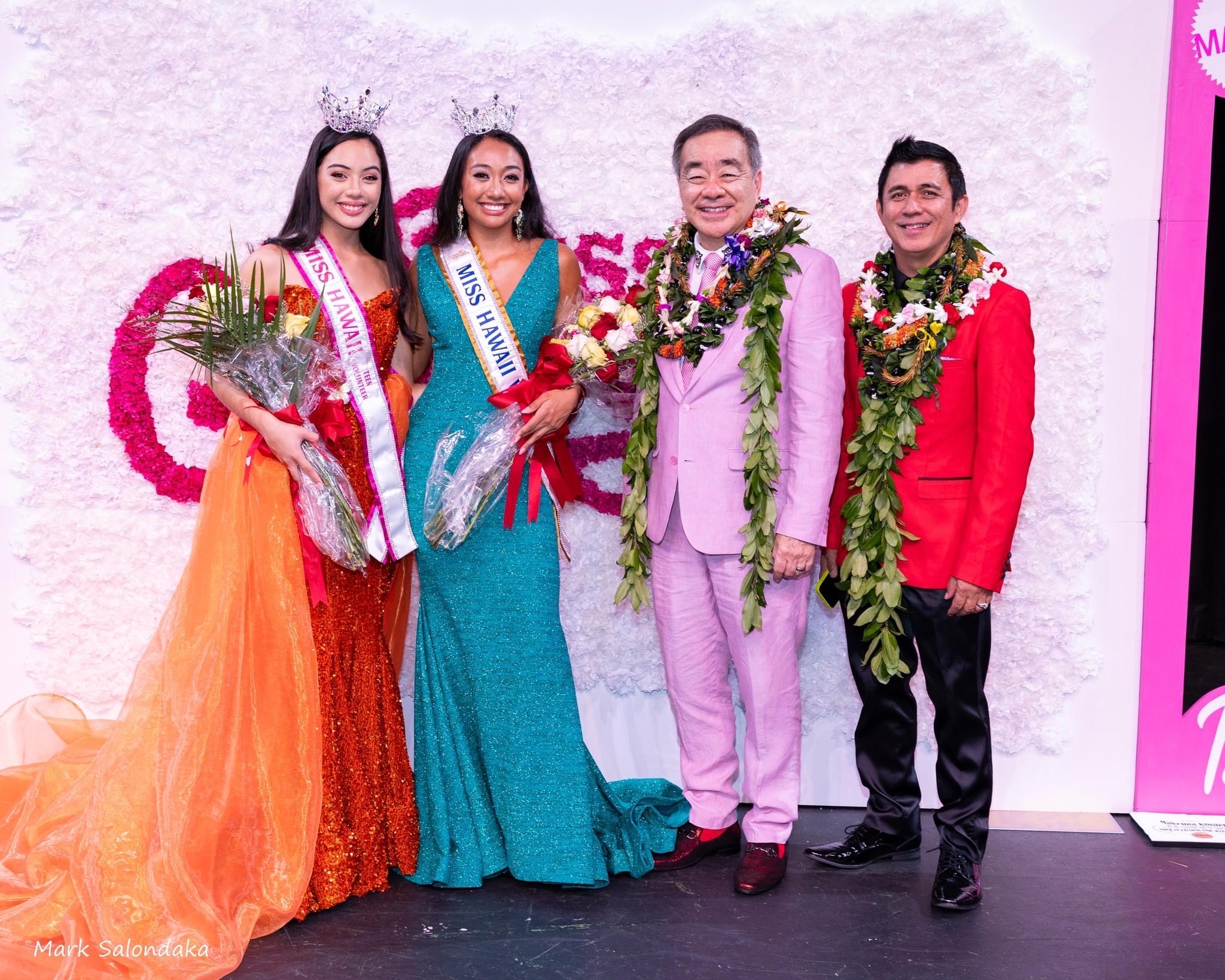 Two women and two men posing for a group photo. The women are wearing pageant gowns and crowns, and holding flowers.
