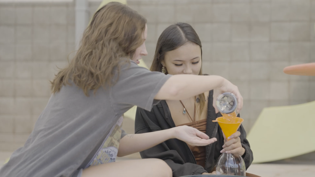 Two students working on a project together, pouring liquid from one bottle into another bottle.