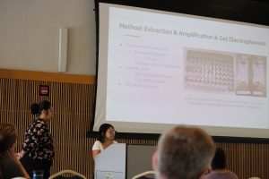 Two students standing at a podium in front of a large screen and before an audience.