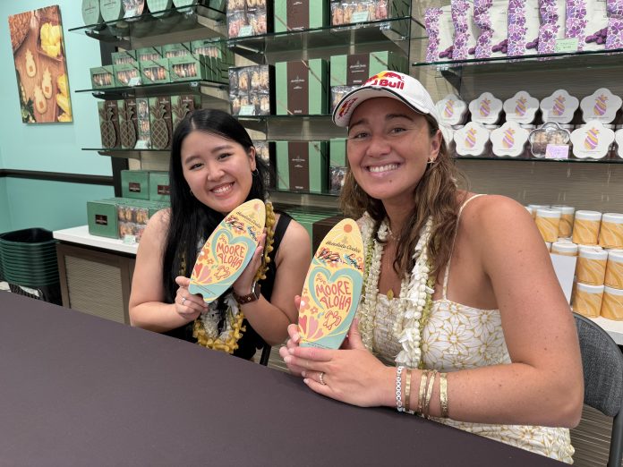 Two women sitting at a table and each holding up surfboard-shaped cookie tins.